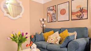 Un salón con un sofá azul, una mesita con un jarrón en forma de casa y un espejo fijado a la pared.