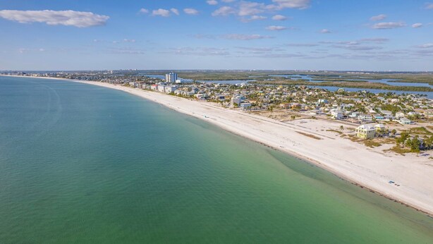 An aerial view of a coastal town with a long sandy beach, clear blue waters, and a dense cluster of buildings along the shore. - Spacious 5-bedroom/4-bathroom house. Less than 1 block to beach! Free Wine! (Fort Myers Beach)