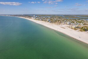 Vue aérienne d’une ville côtière avec une longue plage de sable, des eaux d’un bleu limpide et un ensemble dense de bâtiments le long de la côte.
