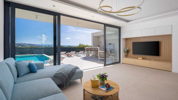 A modern living room with a pool view, a sofa, a coffee table, and a TV mounted on a wooden panel wall. - Luxury Villa Nikandra with Pool (Žrnovnica)
