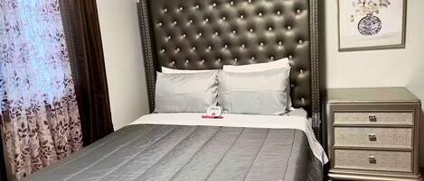 Una cama bien hecha con cabecera capitonada, una mesita de noche con cajones y una obra de arte floral enmarcada en la pared.