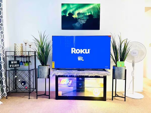 A living room with a large flat-screen TV displaying 'Roku' on the screen, a wine cooler, a small table with a neon sign, and a standing fan.