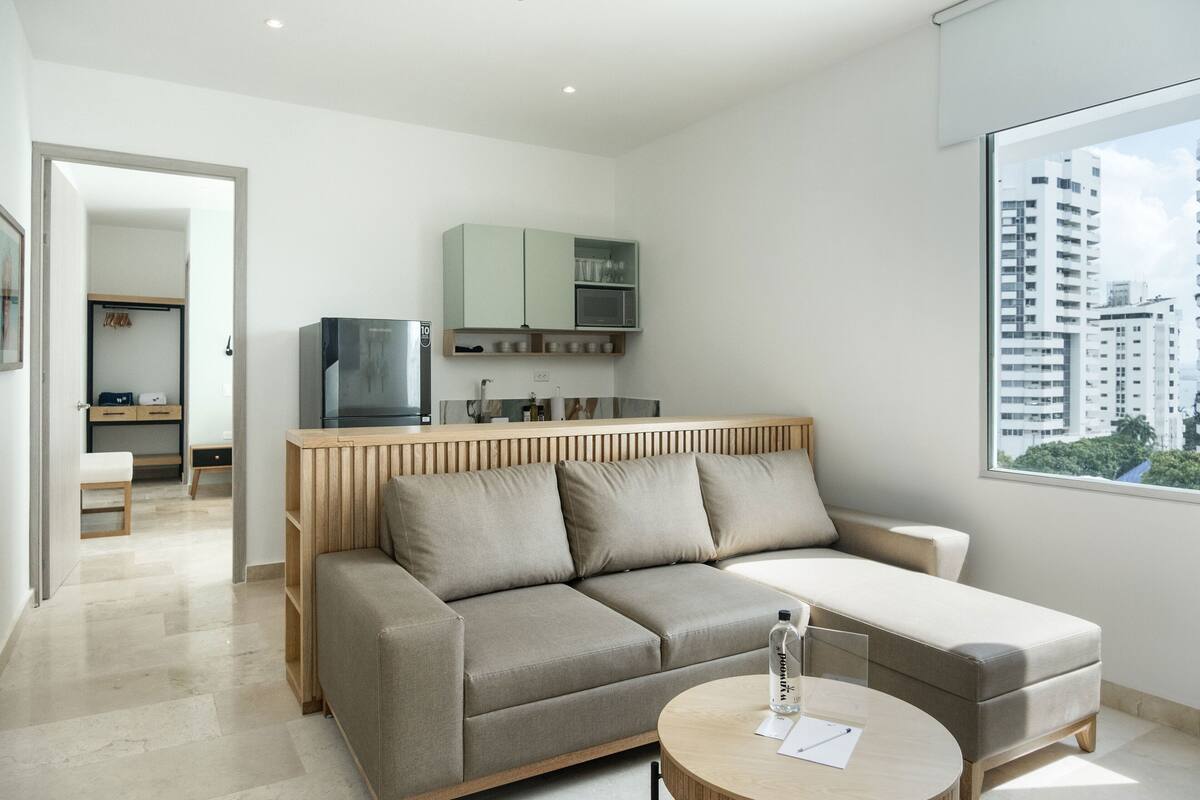 A modern living room with a sofa and a coffee table, a kitchen area with cabinets and a microwave, and a view of a cityscape through a large window.