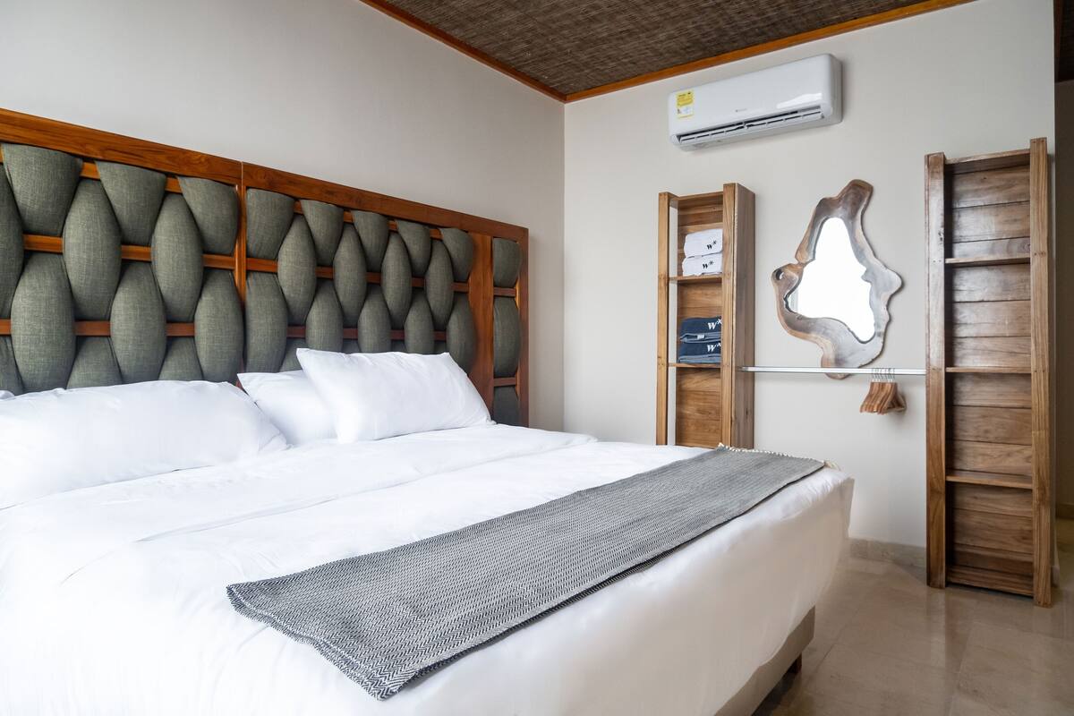A neatly made bed with a patterned throw, a wooden headboard, and a wall-mounted air conditioner.