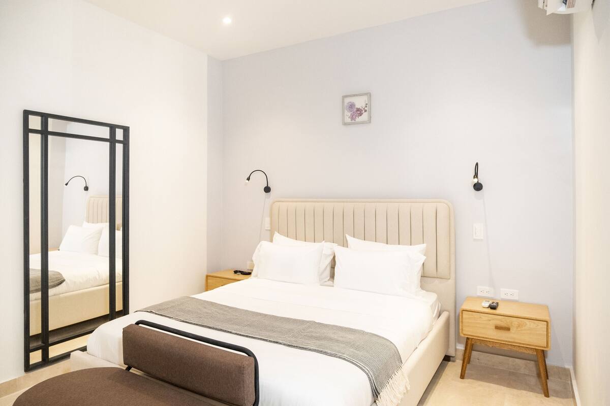 A bedroom with a bed, bedside tables, a nightstand, a mirror, and a wall-mounted light fixture.