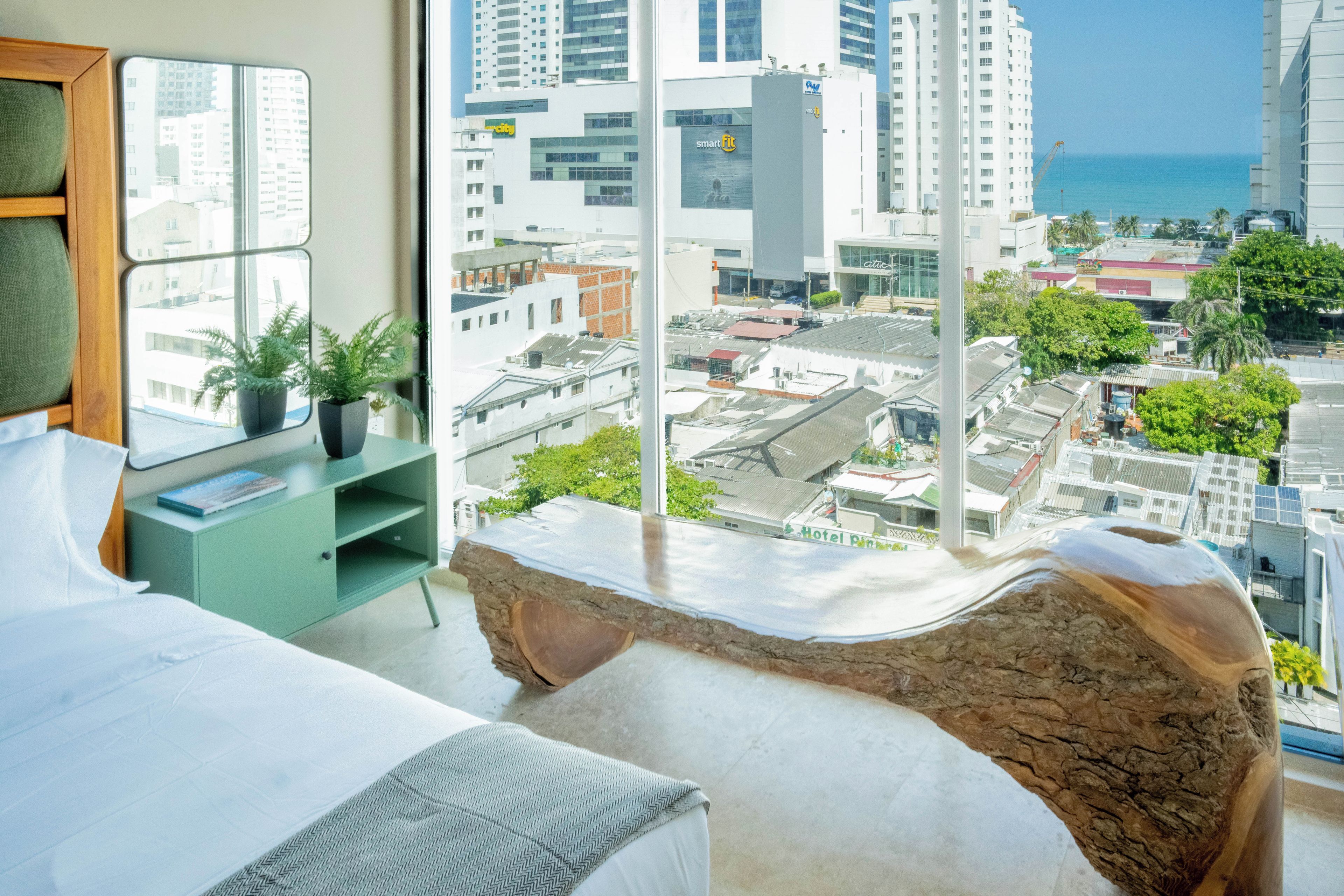 A modern hotel room with a large window, a bed, a green console table, and a view of the city and ocean.