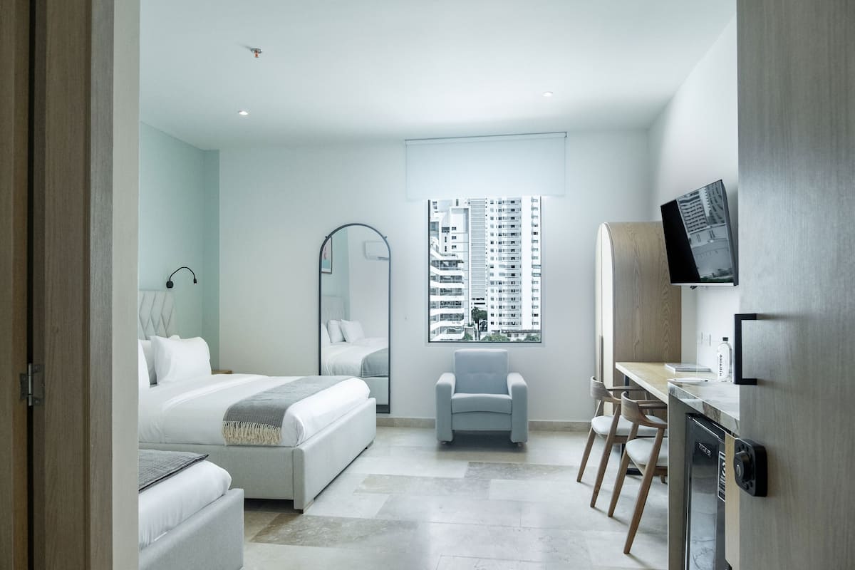A modern hotel room with a bed, a seating area, a dining table, and a view of high-rise buildings.