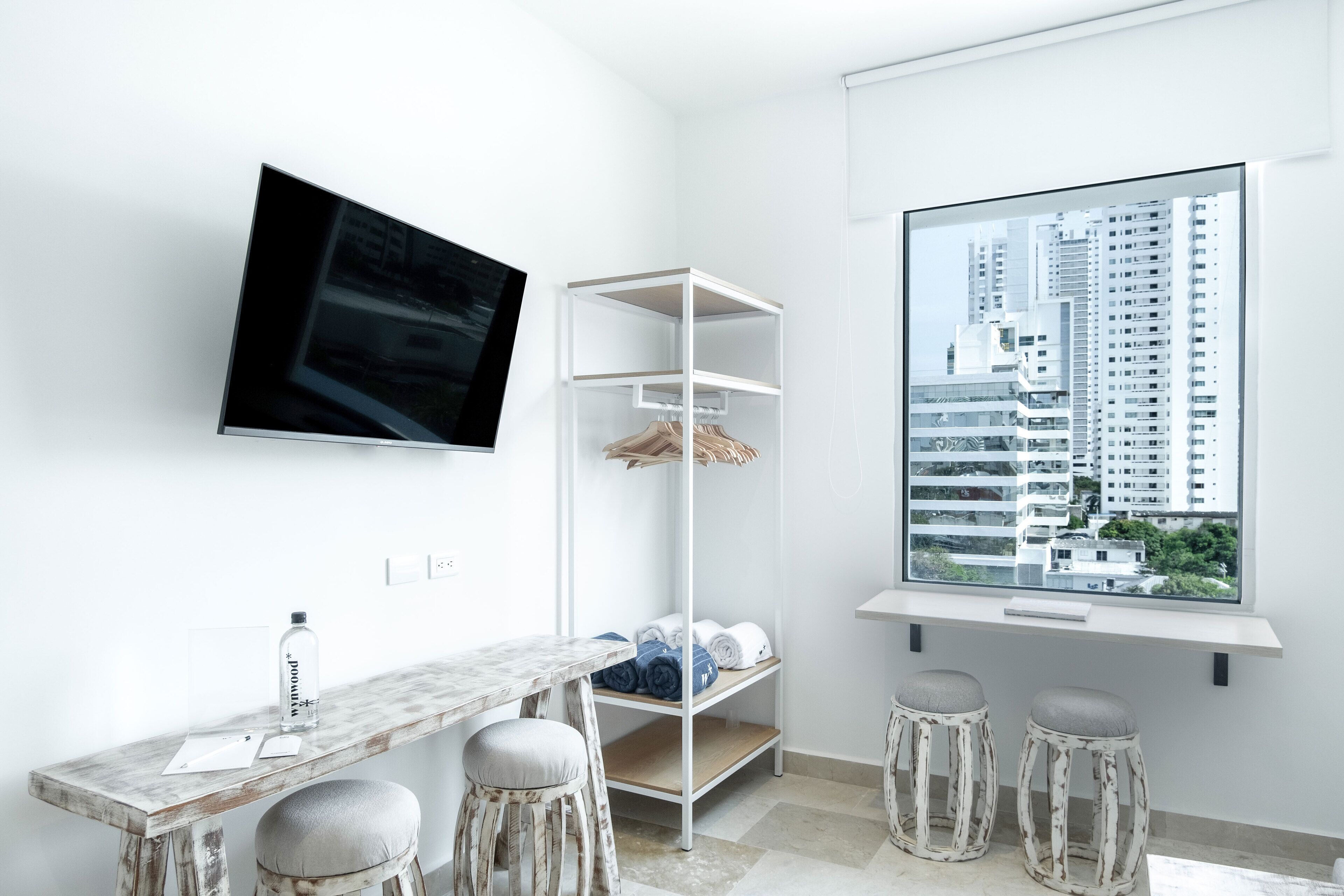 A modern hotel room with a large window, a flat-screen TV, a wooden table with stools, and a white shelving unit.