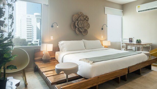 A modern bedroom with a wooden bed, two bedside lamps, a wall-mounted decorative piece, and a view of buildings through a large window. - Mega by Wynwood House (Cartagena)