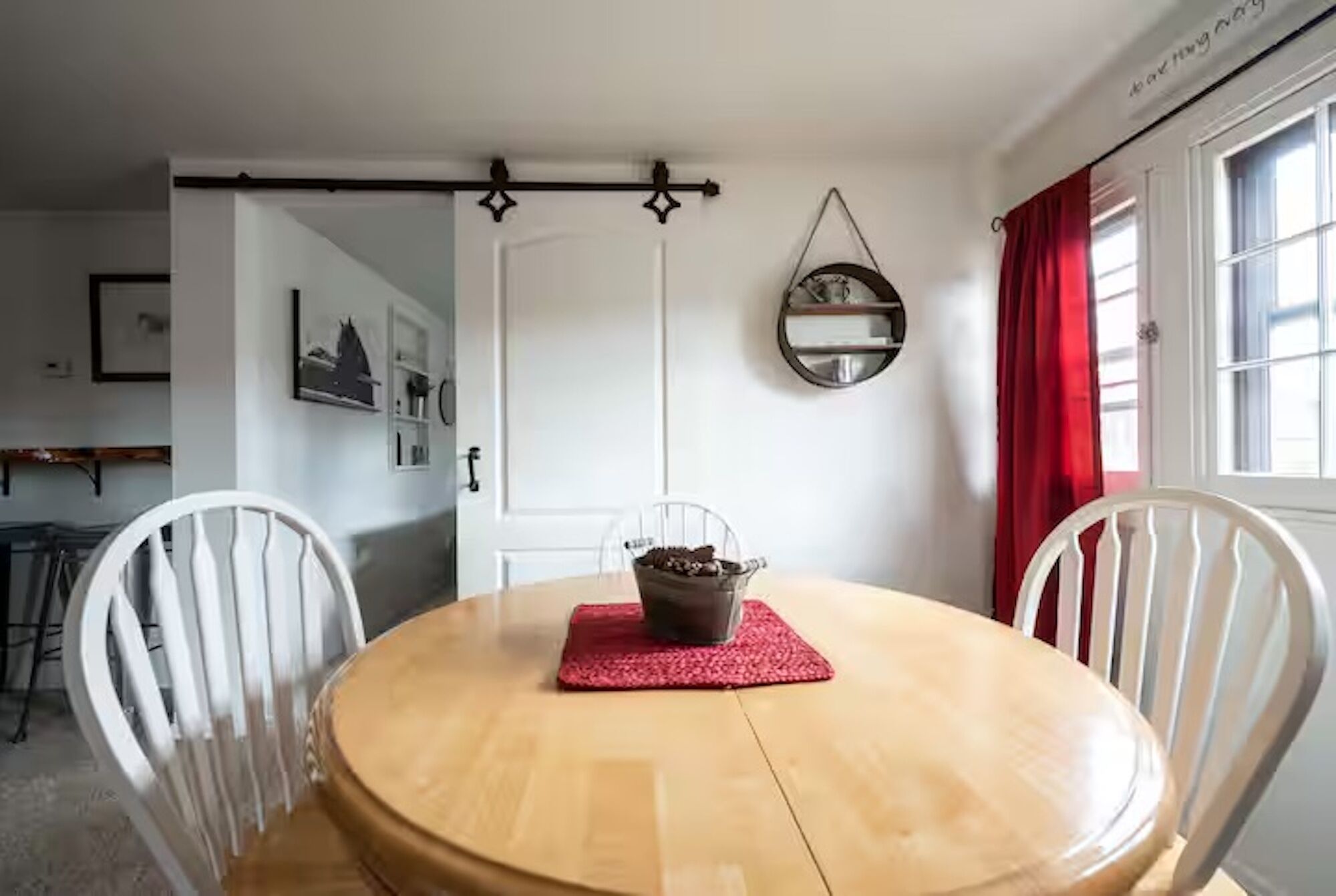 Un comedor con una mesa de madera, dos sillas blancas, un mantel individual rojo y un tazón metálico con chispas de chocolate.