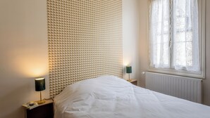 A bedroom with a patterned wall, a bed, two bedside tables with lamps, and a window with lace curtains. - 🏡 Large charming gîte in Fontevraud - Warm winter stays (6 to 12 pers) (Fontevraud-l'Abbaye)