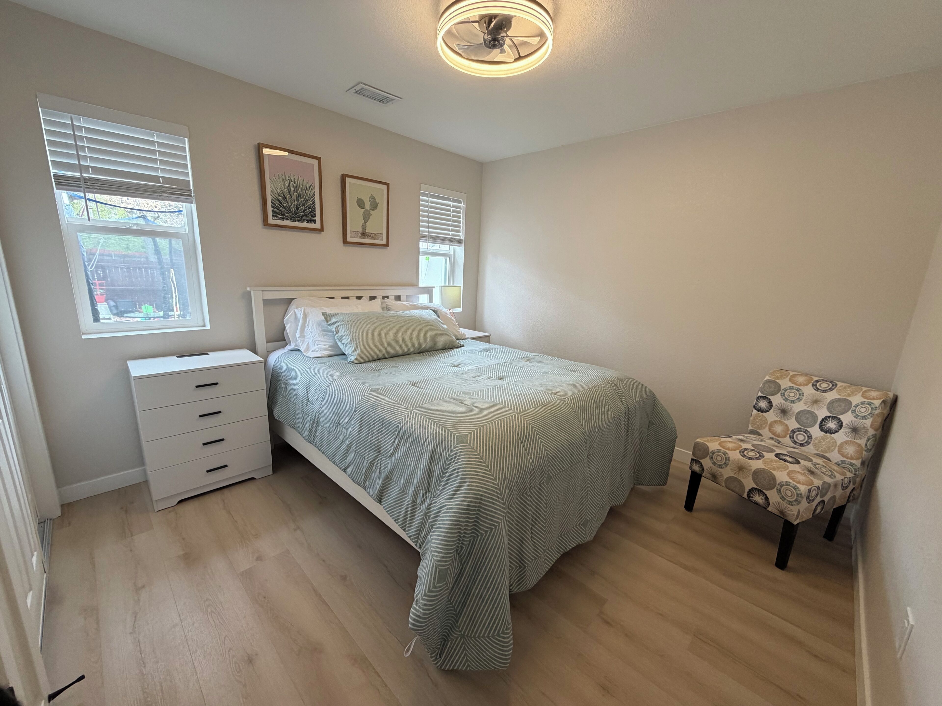 A bedroom with a bed, a nightstand, a chair, and two windows.