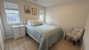 A bedroom with a bed, a nightstand, a chair, and two windows.