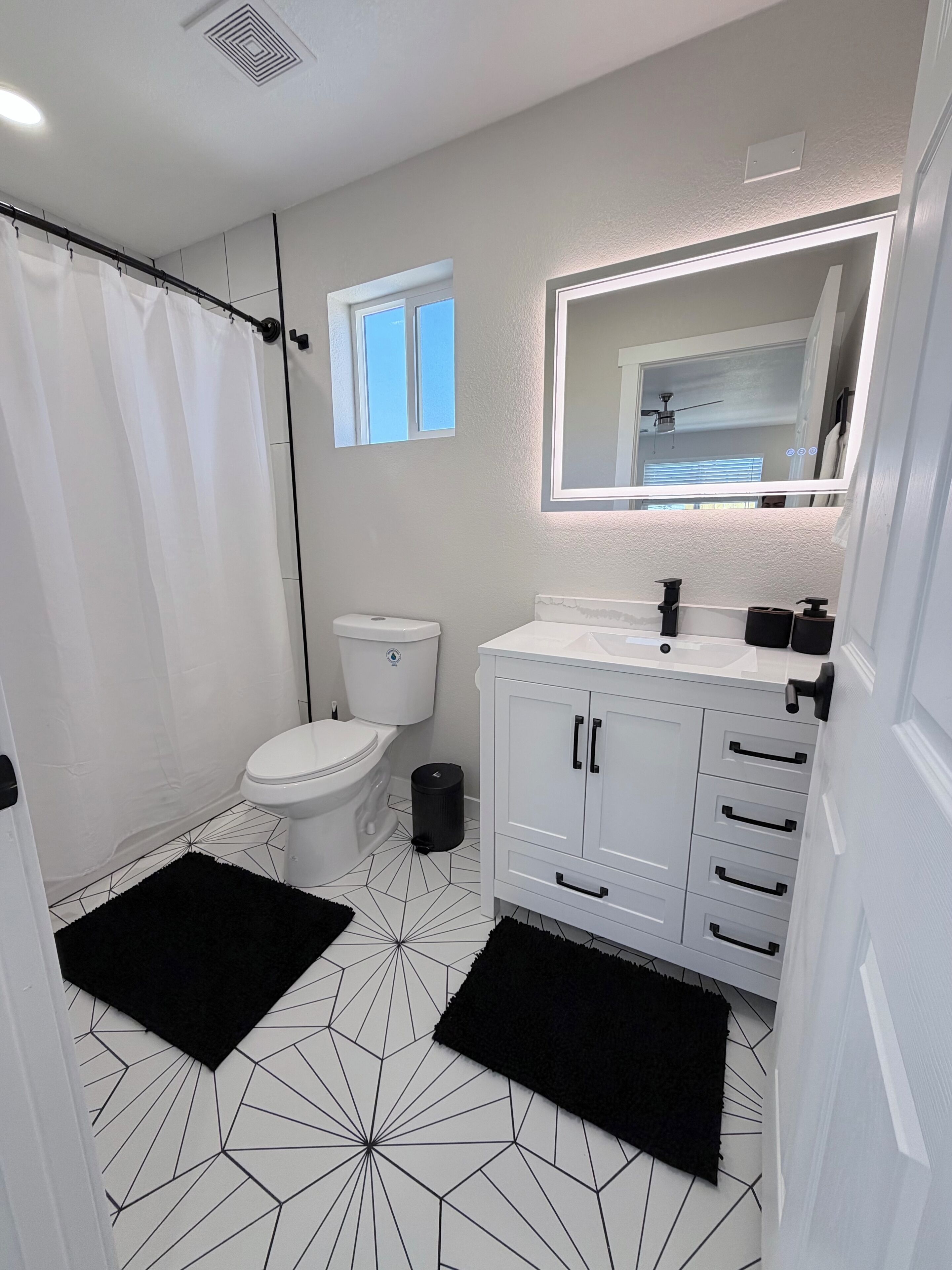 Un bagno con water, lavandino, specchio e tappeti neri su un pavimento in piastrelle geometriche.