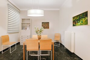 A dining area with a table, chairs, a vase with flowers, a painting on the wall, a radiator, and a bookshelf. - Leda’s Nea Chora Flat - Near Beach & Old Port (Chania)