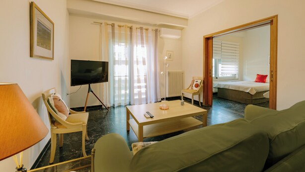 Hotellihuoneessa on vihreä sohva, puinen sohvapöytä, jalustalla oleva televisio, tyynyillä varustettu tuoli, punaisella tyynyllä varustettu sänky ja suuri peili.