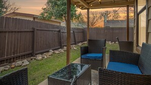 Ein überdachter Patio-Bereich mit Korbmöbeln und einem Glastisch, umgeben von einem Holzzaun und einem Rasen.