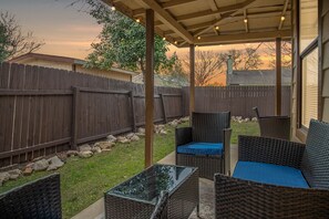 A covered patio area with wicker furniture and a glass table, surrounded by a wooden fence and a grassy yard. - 4B Pet Friendly, Fenced Yard, LacklandAFB, SeaWrld (San Antonio)