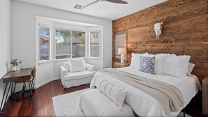 Quarto com cabeceira de madeira, cama com roupa de cama branca, escrivaninha com cadeira e janela com persianas.
