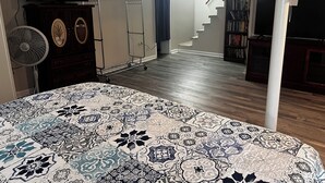 Ein Schlafzimmer mit einem gemusterten Bettbezug, einem Standventilator, einem Schminktisch, einem Fernseher, einem Bücherregal und einer Treppe zu einer weiteren Ebene.
