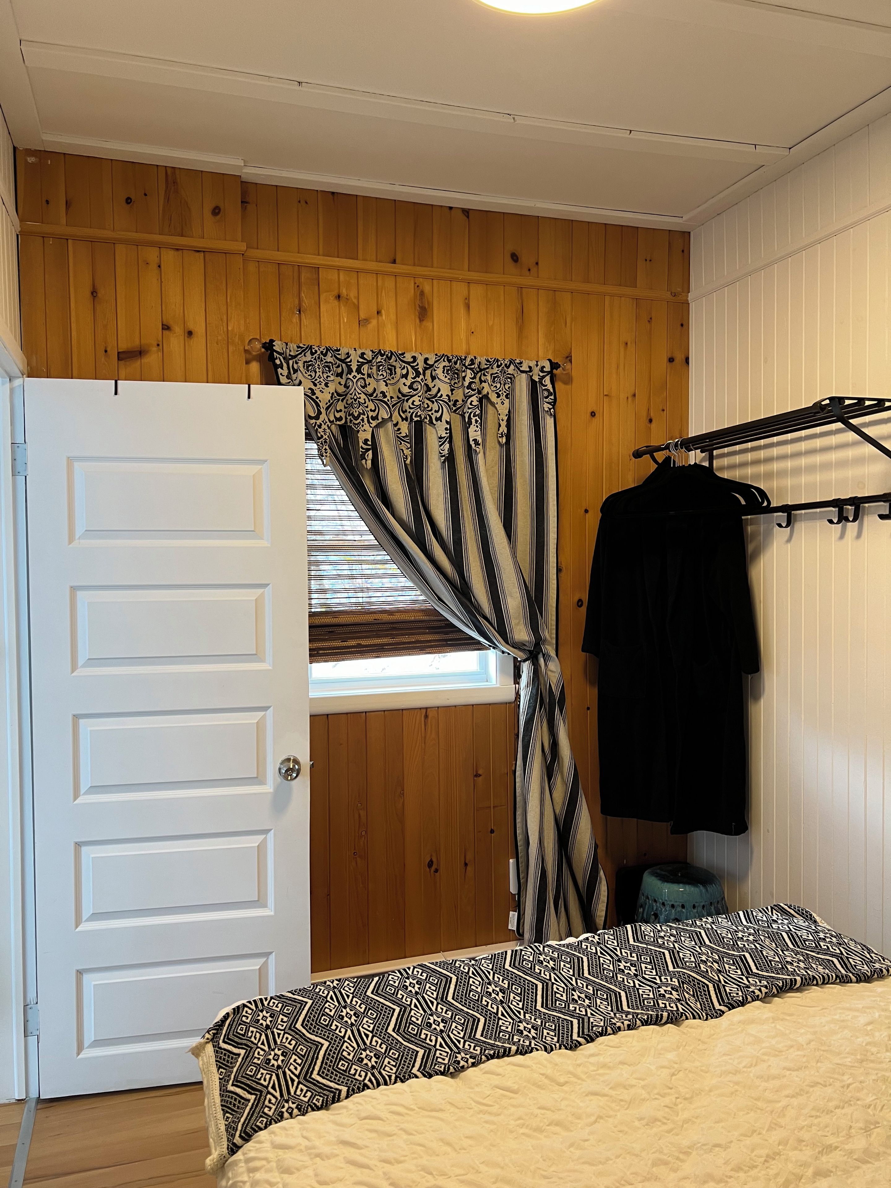 Une chambre avec un plafond en bois, un lit recouvert d’une literie à motifs, une fenêtre avec des rideaux et un portant à vêtements sur lequel est suspendu un vêtement noir.