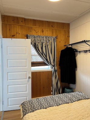 A bedroom with a wooden ceiling, a bed with patterned bedding, a window with curtains, and a clothing rack with a black garment.
