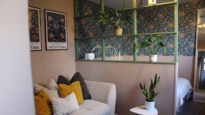 Un saló amb un sofà, coixins, una taula petita amb una planta i una paret amb paper pintat floral i dues obres d'art emmarcades.