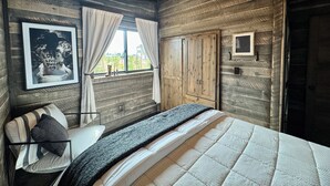 Una habitación de madera con una cama, una silla, un ventanal con cortinas y un cuadro enmarcado en la pared.