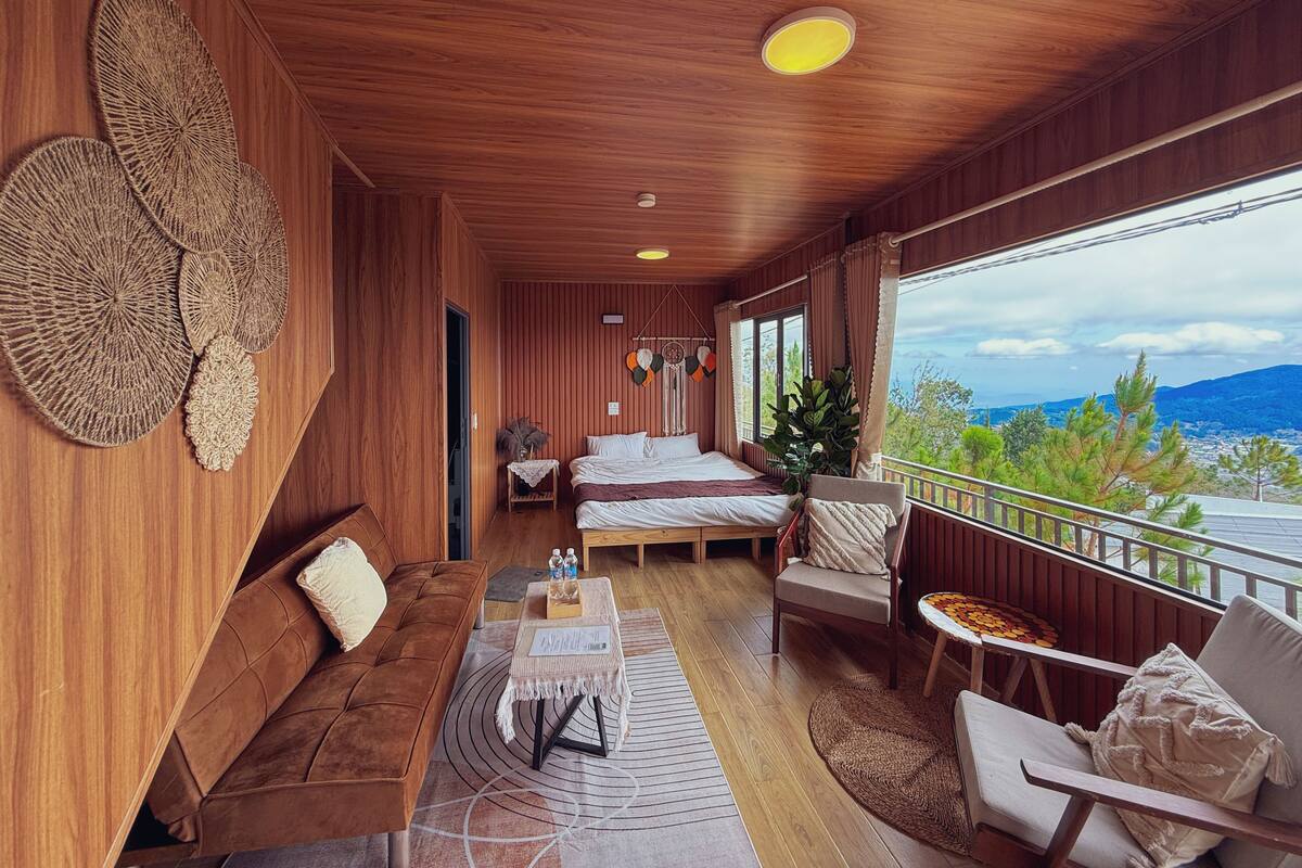 A wooden interior with a bed, sofa, and dining area, a balcony with a view.