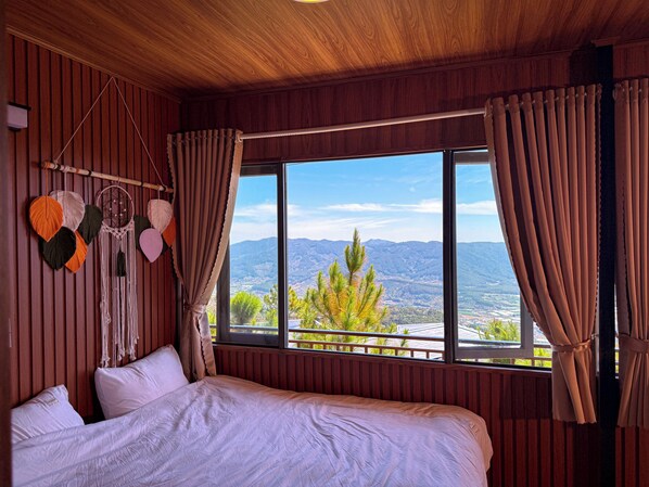 A room with a large bed, wooden panel walls, a view of mountains, and a macramé wall hanging. - Rio Skyhouse (Xuan Truong - Da Lat)