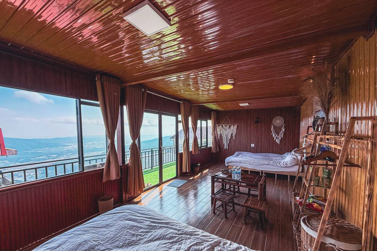 A room with a large bed, a smaller bed, a desk, and a chair. There are windows with a view of the sea and mountains.