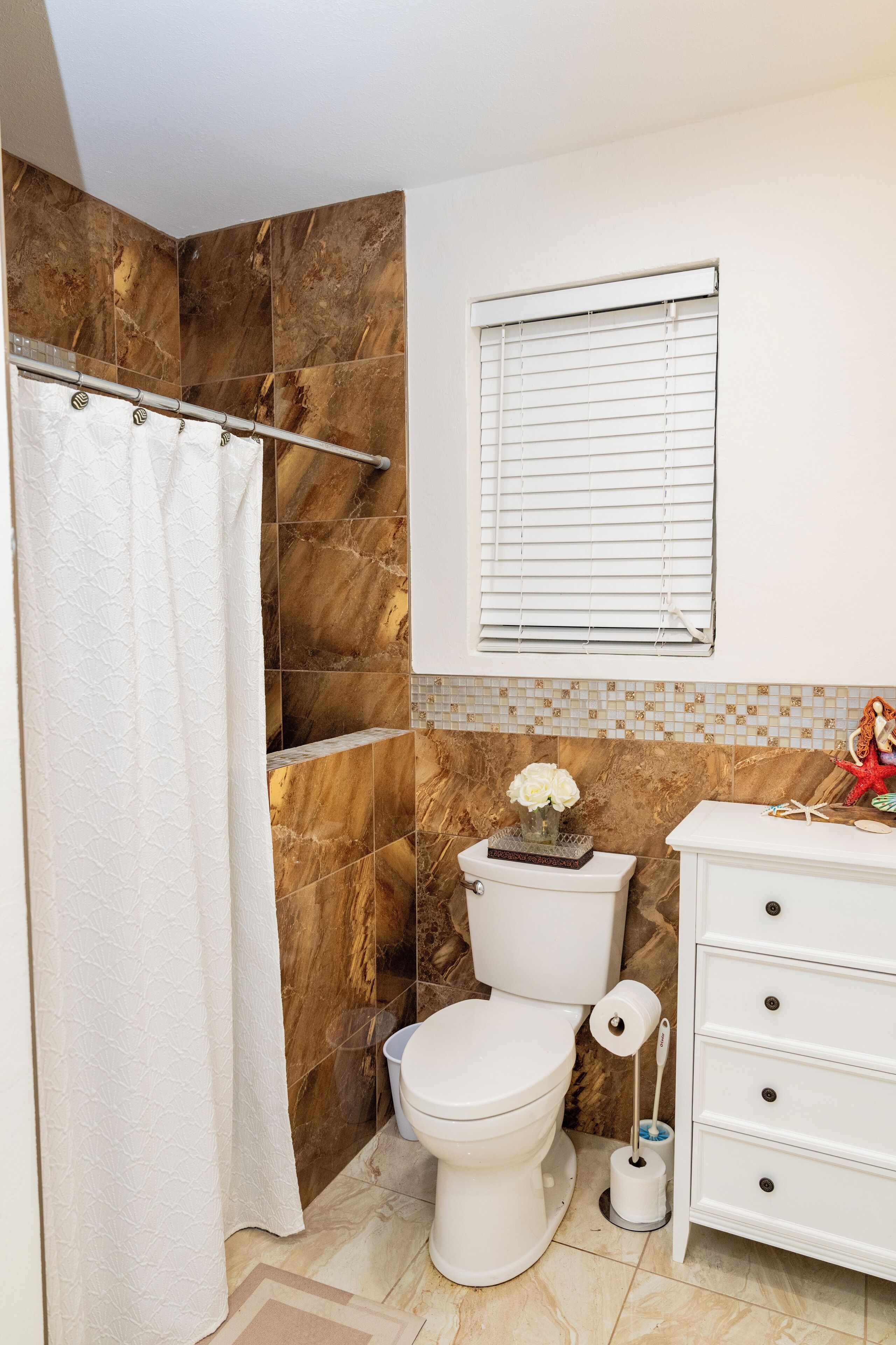 A bathroom with a shower curtain, a toilet, a bidet, a vanity with drawers, and a window with blinds.