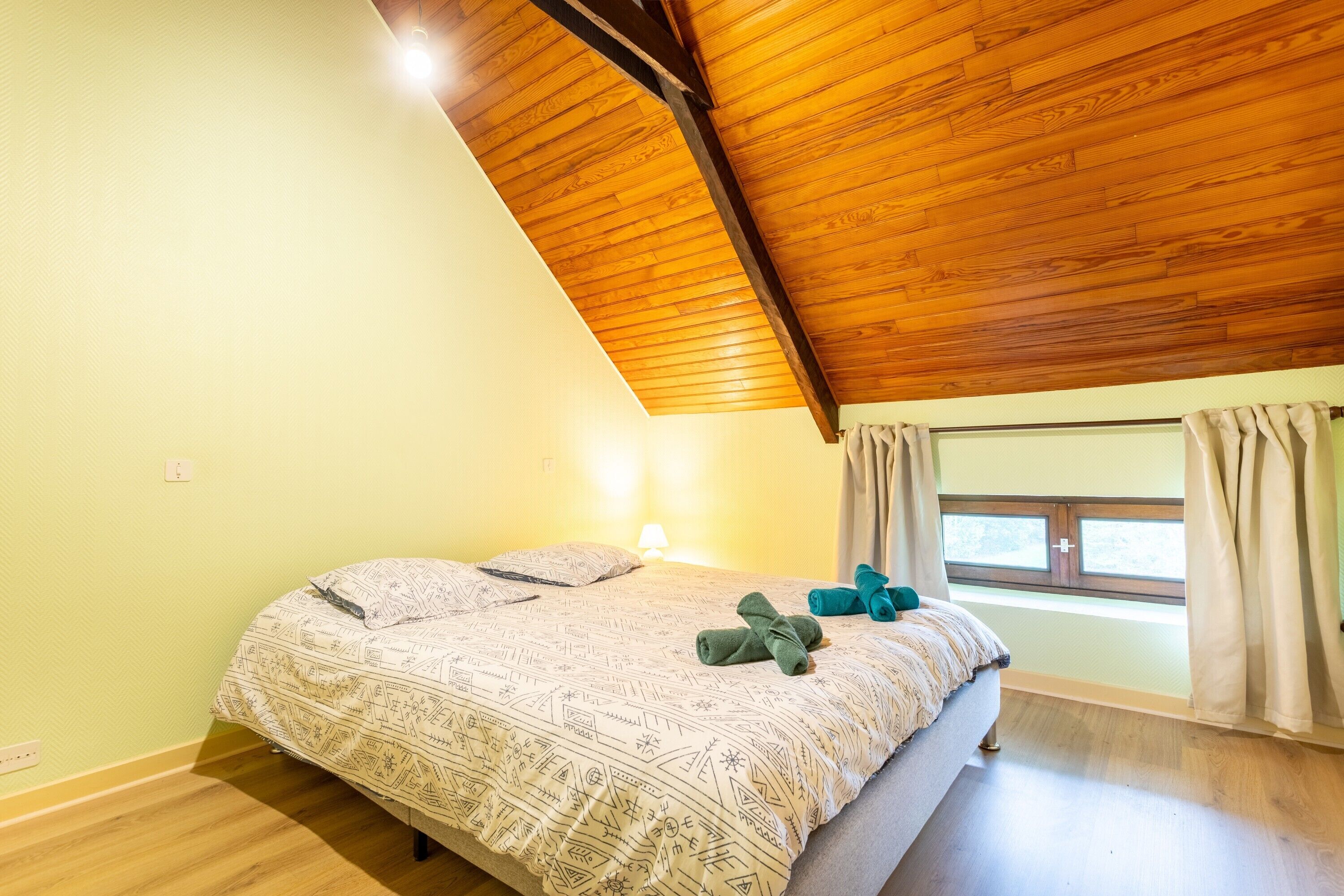 Ein Schlafzimmer mit schrägverdeckter Holzbalkendecke, einem Bett mit gemusterten Betttextilien, einem Fenster mit Vorhängen und einer Wandlampe.