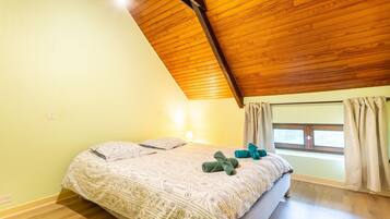 Ein Schlafzimmer mit schrägverdeckter Holzbalkendecke, einem Bett mit gemusterten Betttextilien, einem Fenster mit Vorhängen und einer Wandlampe.