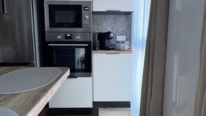 A modern kitchen with stainless steel appliances, a built-in oven, and a microwave. There is a wooden table with a black mat, and a window with curtains. - The white nest in the heart of the Jura mountains (St Laurent, en Grandvaux)