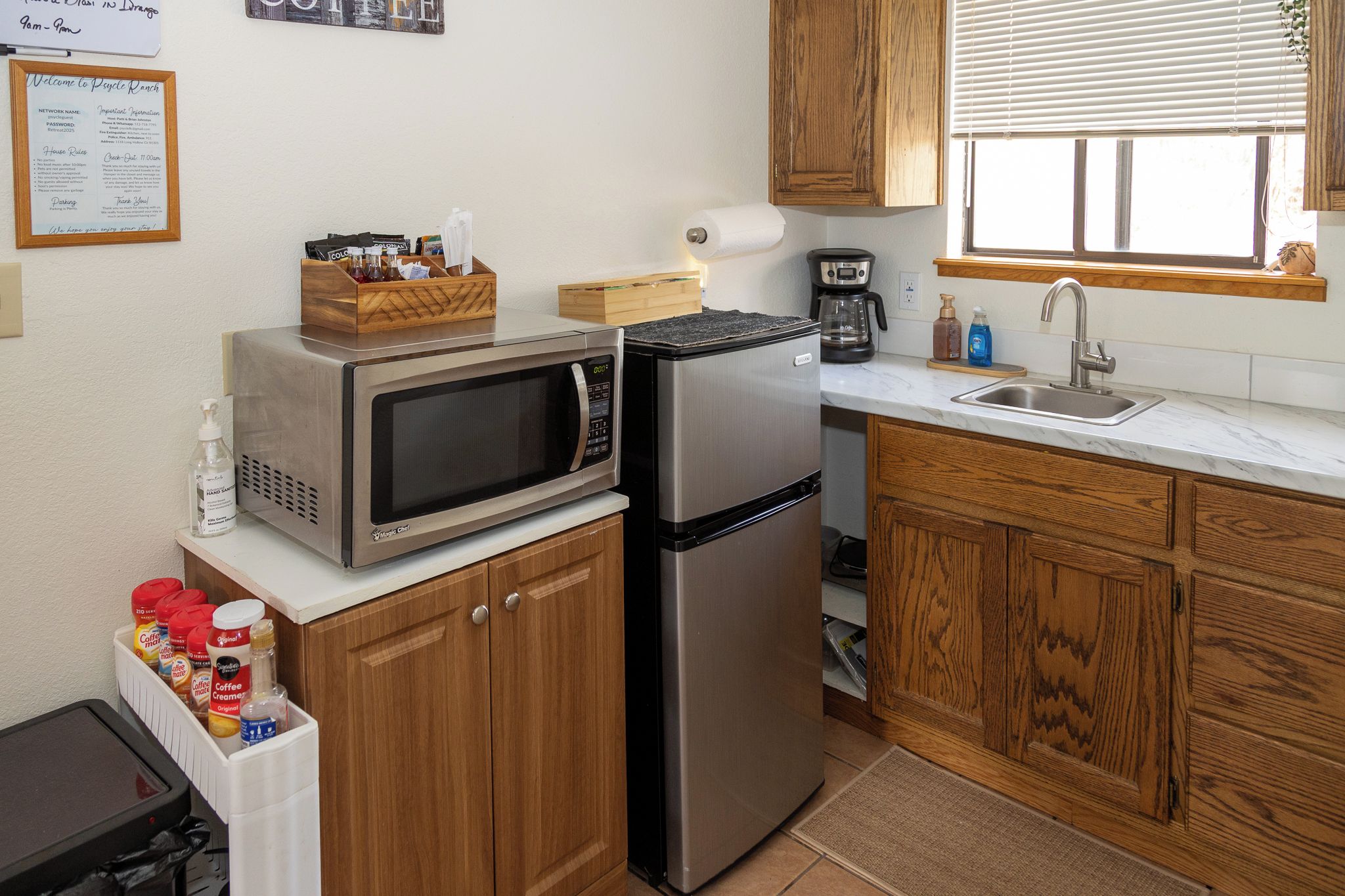 A compact kitchen with a microwave, refrigerator, and wooden cabinets. There is a coffee maker and a sink with a window above it.