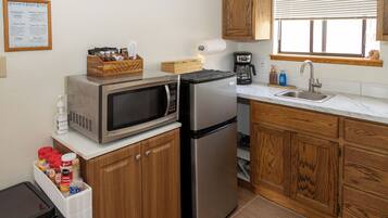 A compact kitchen with a microwave, refrigerator, and wooden cabinets. There is a coffee maker and a sink with a window above it.