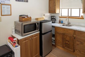 A compact kitchen with a microwave, refrigerator, and wooden cabinets. There is a coffee maker and a sink with a window above it.