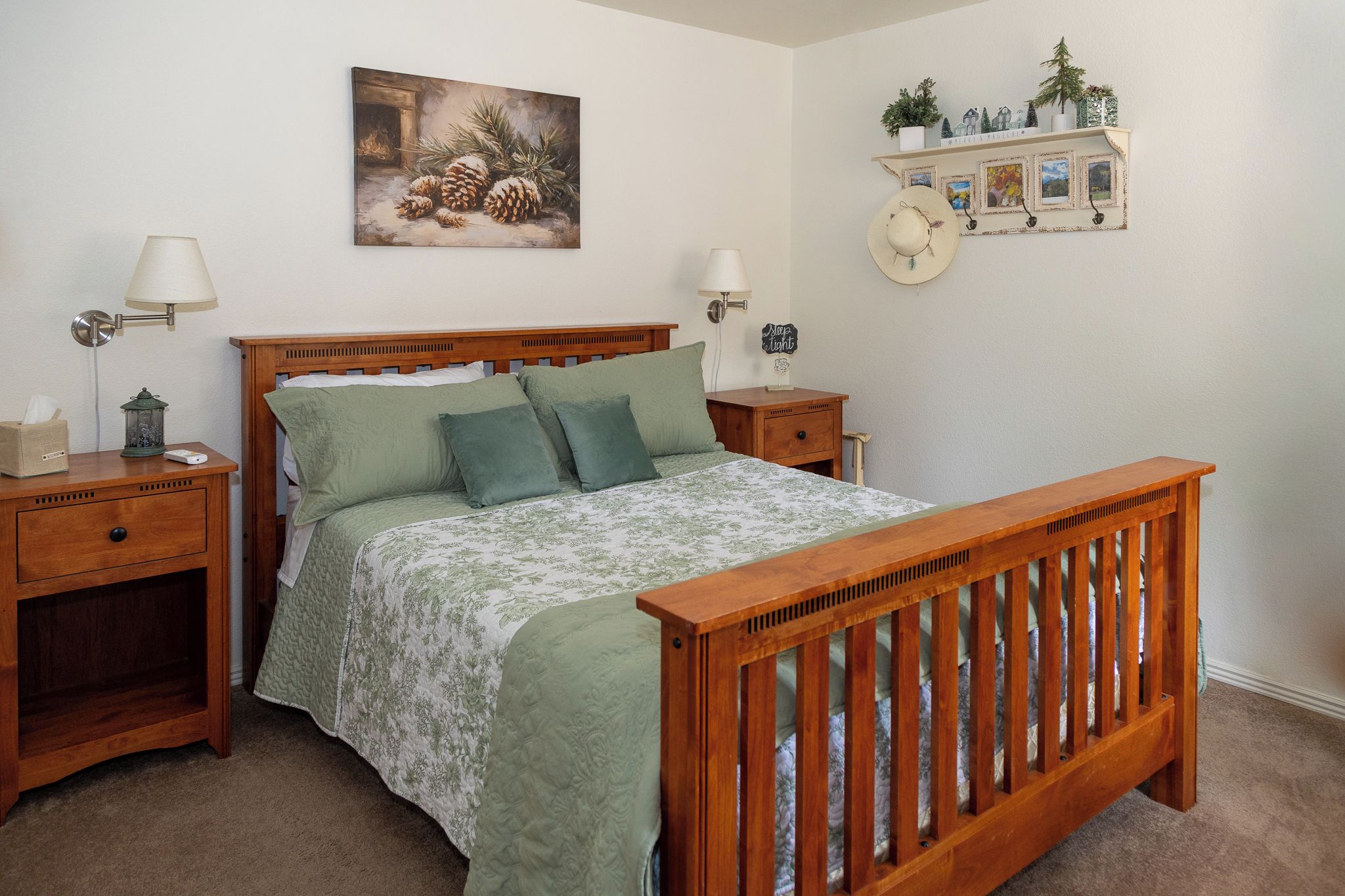A bedroom with a wooden bed, two bedside tables, a nightstand, a lamp, a wall-mounted shelf with decorative items, and a framed picture above the bed.