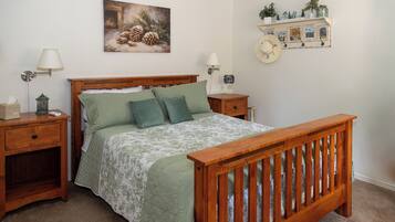 A bedroom with a wooden bed, two bedside tables, a nightstand, a lamp, a wall-mounted shelf with decorative items, and a framed picture above the bed.