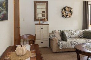 A cozy living room with a wooden chair, a sofa with patterned cushions, a small table with a woven basket, and a decorative wreath on the wall.