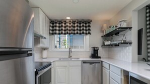 Una cocina moderna con electrodomésticos de acero inoxidable, armarios blancos, un ventanal con un toldo a cuadros y una cafetera sobre la encimera.