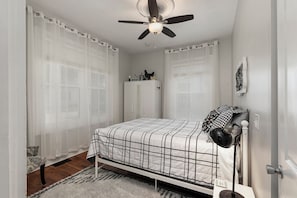 A bedroom with a bed, a ceiling fan, a bedside lamp, and a window with sheer curtains.