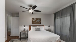 Un dormitorio con cama, dos mesitas de noche, un ventilador de techo, una puerta, un cuadro y una ventana con cortinas.