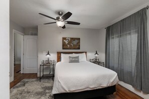 A bedroom with a bed, two bedside tables, a ceiling fan, a door, a painting, and a window with curtains. - Bright & Beautiful Turn-of-the-Century Modern Home (Springfield)