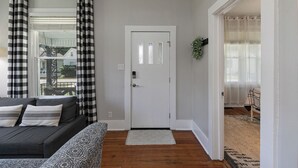 Una sala de estar con un sofá, un reposapiés estampado, una puerta blanca y vistas a través de una ventana con cortinas a cuadros.
