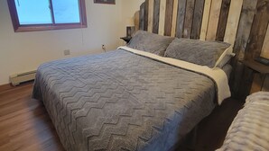 Een slaapkamer met een houten hoofdbord, een groot bed met een grijze deken, een raam met uitzicht op sneeuw, en een klein nachtkastje met een lamp.