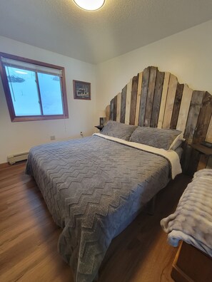 Ein Schlafzimmer mit einem hölzernen Kopfteil, einem großen Bett mit grauer Bettdecke, einem Fenster mit Blick auf Schnee und einem kleinen Nachttisch mit Lampe.