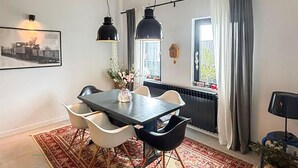 Ein Essbereich mit Tisch, Stühlen und hängenden Lampen. Ein Fenster mit Vorhängen und ein Heizkörper darunter.