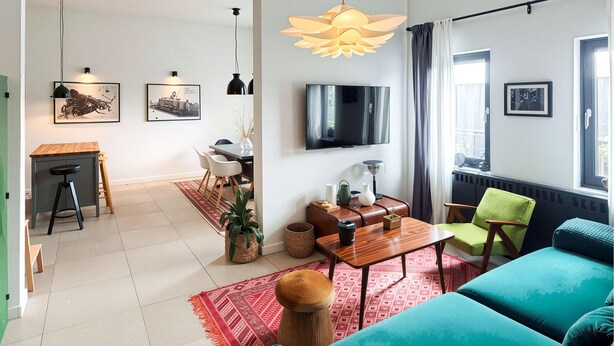 Ein modernes Wohnzimmer mit einer Couch, einem Couchtisch und einem an der Wand montierten Fernseher. Es gibt einen Essbereich mit Tisch und Stühlen sowie eine Küchenzeile mit einem Herd.